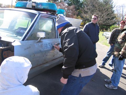 2001 Ford Crown Victoria - A man writes 'CAR 54' on the side of a light blue sedan equipped with blue emergency lights.