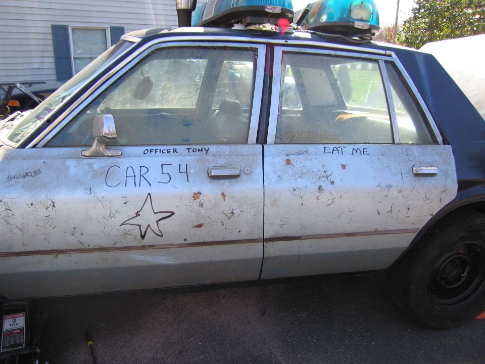 Side view of a beat-up police car with 'CAR 54' and 'OFFICER TONY' written on the door