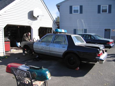 2001 Ford Crown Victoria - A police car is undergoing repairs in a driveway, with a second vehicle parked behind it.