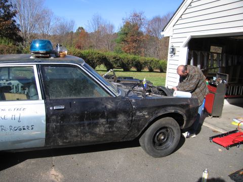 2001 Ford Crown Victoria - A man is working on the engine of a weathered car that has a police light on its roof and custom text on its side.