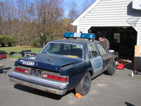 2001 Ford Crown Victoria - A beat-up police car sits in a driveway next to an open garage.