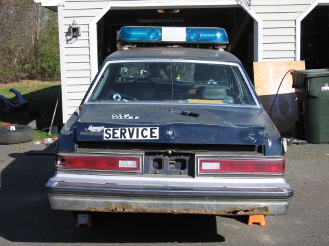 2001 Ford Crown Victoria - Rear view of a dark blue police car with a light bar, parked in front of a garage.