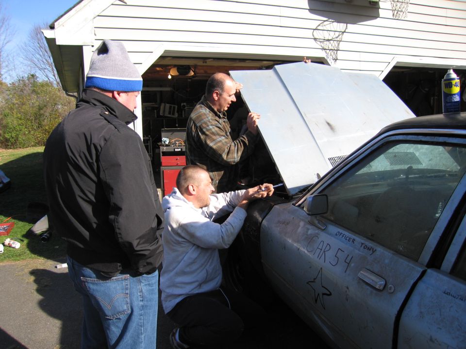 Side view of a weathered car with 'CAR 54' written on the door, as three men work on the engine