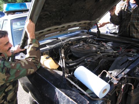 2001 Ford Crown Victoria - Two men examine the engine compartment of a dark-colored vehicle, with a police car visible behind them.