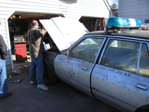 2001 Ford Crown Victoria - A police car with its hood open is being worked on by two men in front of a garage.