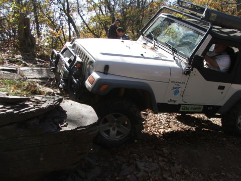 1996 Ford Bronco - A white Jeep Wrangler navigates a challenging rocky trail with trees in the background.
