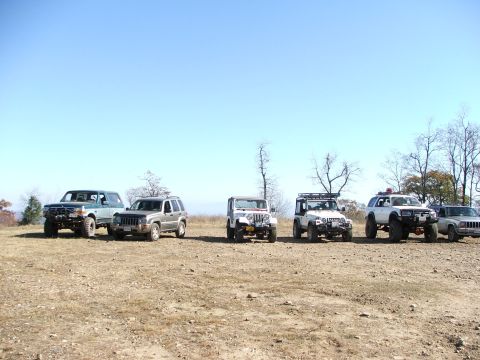 1996 Ford Bronco - A group of off-road vehicles are parked in a line on a dry, grassy area with trees in the background.