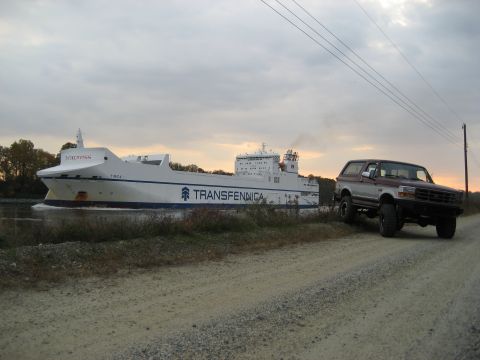 1996 Ford Bronco - 1996 Ford Bronco parked on a dirt road beside a large cargo ship sailing on a canal.