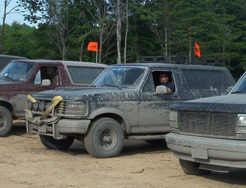 Muddy 1993 Ford Bronco with off-road lights and a driver visible. Another Bronco is to its left, and part of a third vehicle is on the right