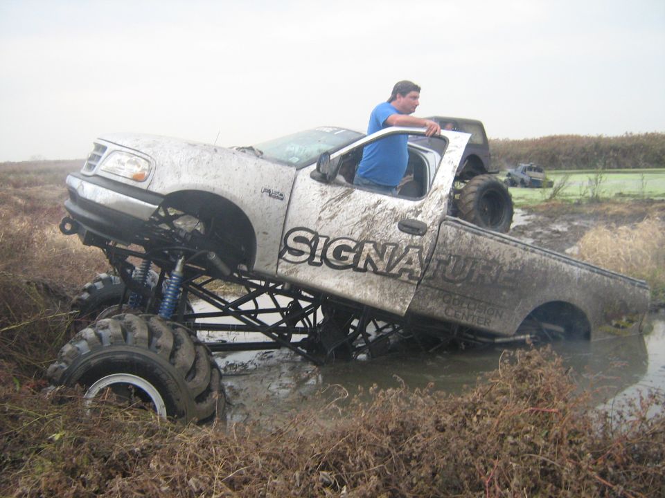 Lifted pickup truck with 'SIGNATURE' on the side stuck in mud, driver leaning out window. Another vehicle visible in background