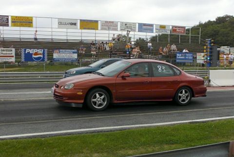 1997 Ford Taurus - Two cars are side-by-side on a drag strip, with spectators in the bleachers behind them.