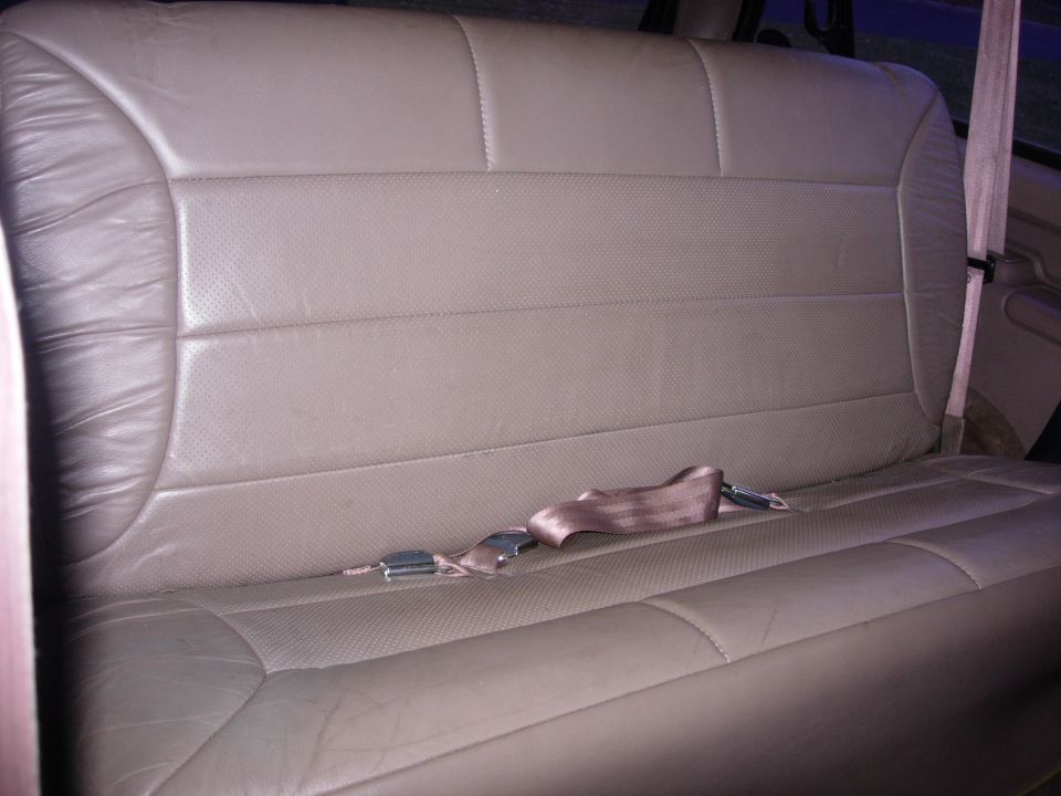 Tan leather rear bench seat with a buckled seatbelt across it. The seat has perforated inserts and visible stitching