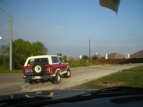 1984 Ford Bronco - A red and white SUV drives on a dirt road with houses in the background.