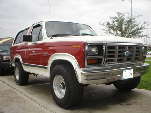 1984 Ford Bronco - This two-tone red and white Ford Bronco is seen from a 3/4 front angle, with another vehicle partially visible to its left.