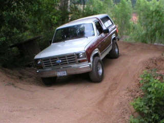 1984 Ford Bronco - A two-tone Ford Bronco navigates a dirt path surrounded by trees.
