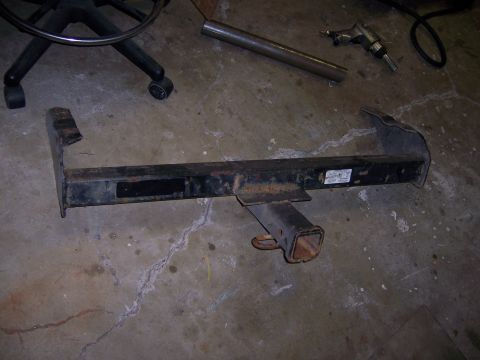 1991 Ford Bronco - A black trailer hitch lies on a concrete floor, with a metal pipe and part of an office chair visible.