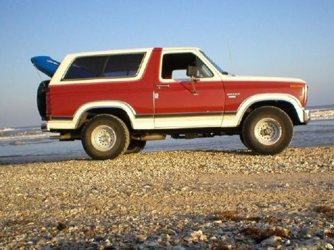 1984 Ford Bronco - This red and white Ford Bronco is parked on a rocky beach near the ocean.
