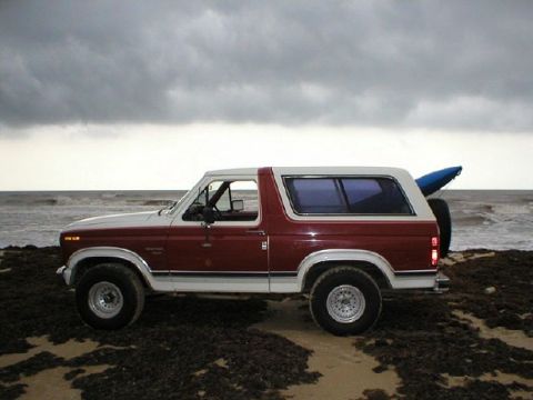 1984 Ford Bronco - A two-tone Ford Bronco sits on the shore, ready for an adventure with a kayak secured to its rear.
