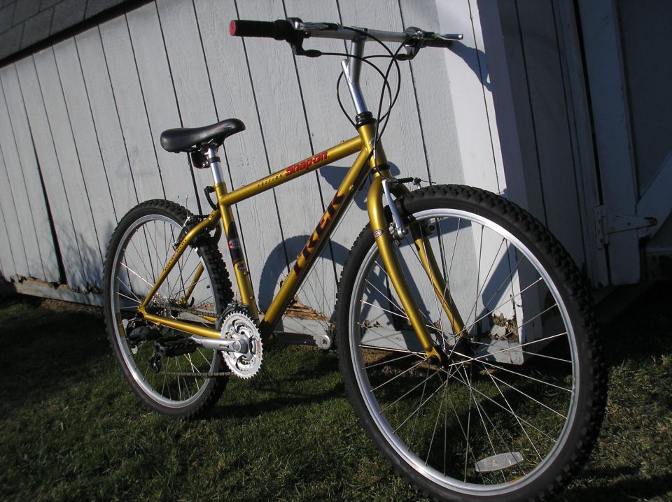 Gold Trek mountain bike with red handlebar grips leaning against a light-colored wall