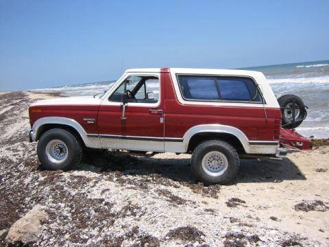 1984 Ford Bronco - A red and white 1984 Ford Bronco is parked on a sandy beach with the ocean in the background.