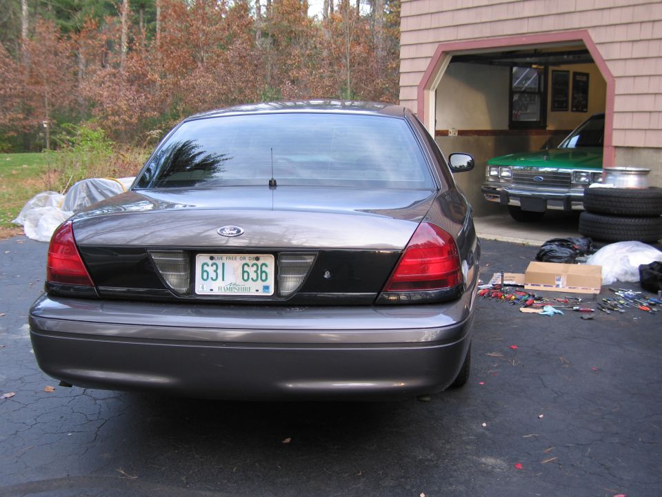Rear view of a gray Ford Crown Victoria parked on asphalt, with a green vehicle visible in a garage behind it