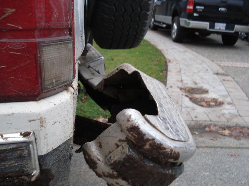 Truck's bent rear bumper and spare tire, red taillight, and white/black body panel. Another black truck is visible in the background