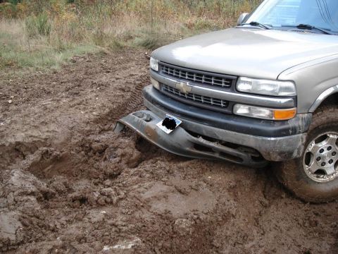 1986 Ford Bronco - A silver Chevrolet pickup truck is stuck in thick mud, with its front bumper hanging loose.