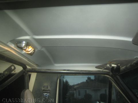 1990 Ford Bronco - vehicle interior view of the headliner, roll-cage bar, lit dome light and rear window, with houses visible through the glass.