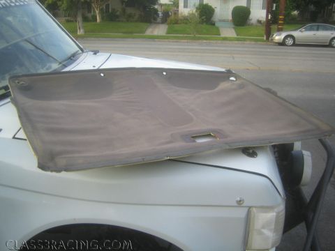 1990 Ford Bronco - removed fabric headliner panel rests propped on the hood of a white truck parked at a residential curb with a car passing behind.