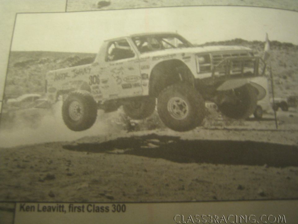 Sepia photo of a white off-road race truck number 300 jumping over dirt in the desert