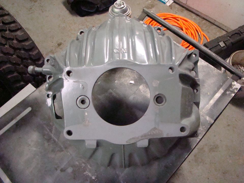 Gray metal automotive part with a circular opening and bolt holes, resting on a metal surface. Orange extension cord and tire are visible in the background
