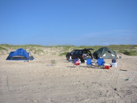 1984 Ford Bronco - A campsite is set up on a sandy beach with a dark SUV, two tents, and chairs under a clear blue sky.