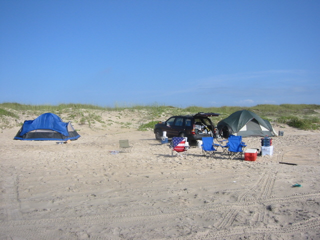 Dark SUV with open trunk parked on a sandy beach next to two tents and camping chairs