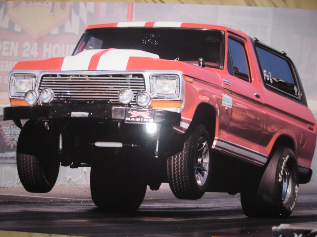 Orange Ford Bronco with white stripes doing a wheelstand, front three-quarter view