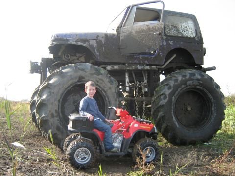 1990 Ford Bronco - A child on a small ATV poses in front of a colossal, mud-splattered monster truck.