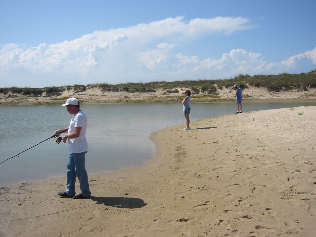 Man in white cap and blue jeans fishes on beach; two other people fish in distance