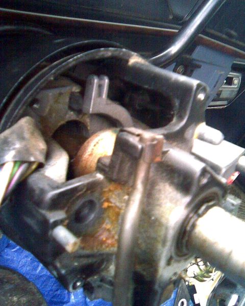 1989 Ford Bronco - Tight close-up of a steering column torn down inside a vehicle, showing the cast tilt housing, brass shift tube, turn-signal switch bracket…
