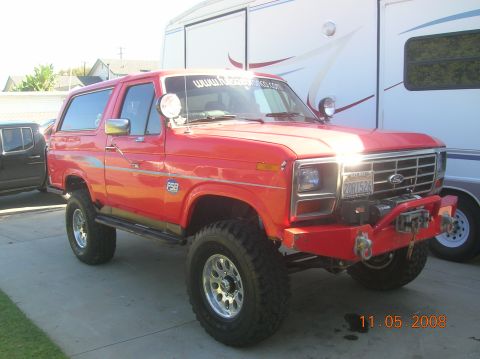 1985 Ford Bronco - A red Ford Bronco is parked in front of a large white trailer on a sunny day.