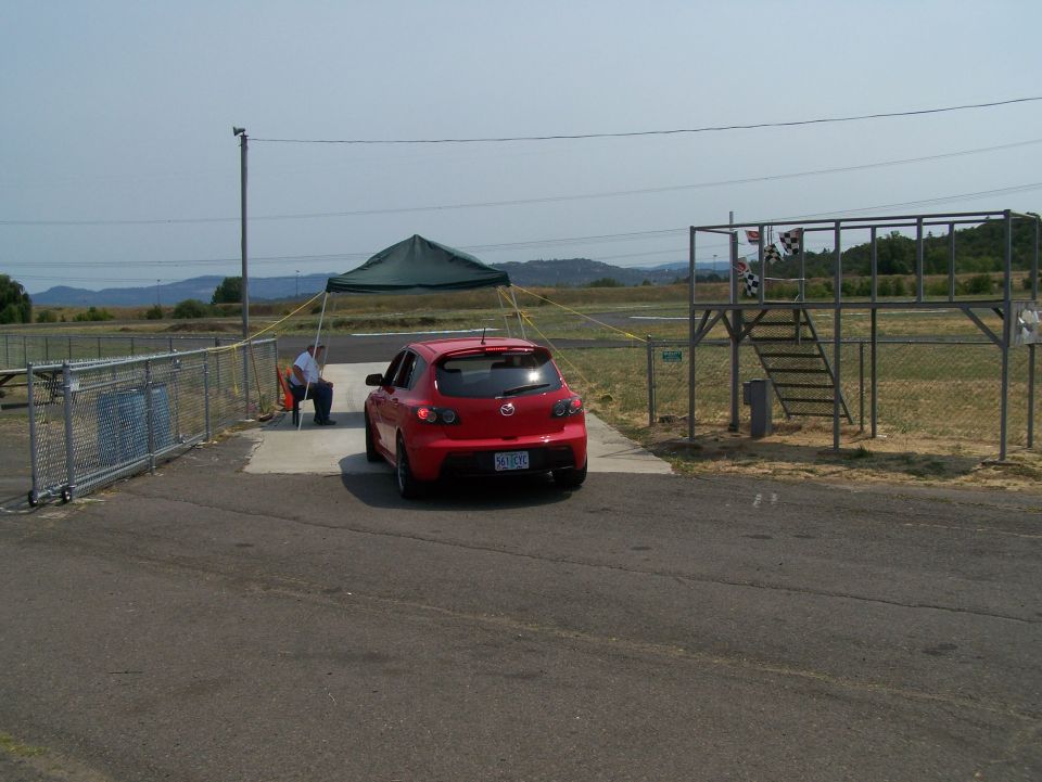 Red 2007 Mazda 3 hatchback parked near a racetrack structure with checkered flags