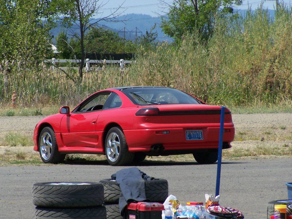 Red coupe with black roof, rear view, parked on dirt and asphalt.