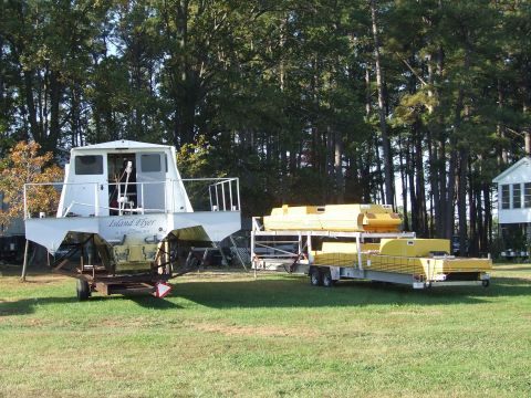 1976 Ford F-150 - A white houseboat named 'Island Flyer' sits on a trailer next to two trailers loaded with yellow pontoons.