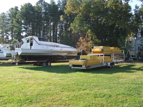 1976 Ford F-150 - A white catamaran boat and a yellow cargo trailer sit on a grassy area under trees.