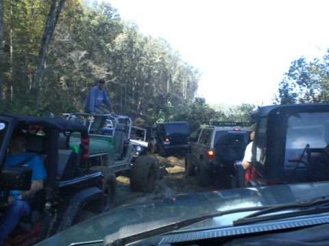 1989 Ford Bronco - Several off-road vehicles navigate a challenging muddy trail surrounded by trees.