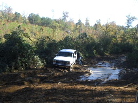 1989 Ford Bronco - A white SUV is stuck in a muddy off-road trail, with a person attempting to assist.