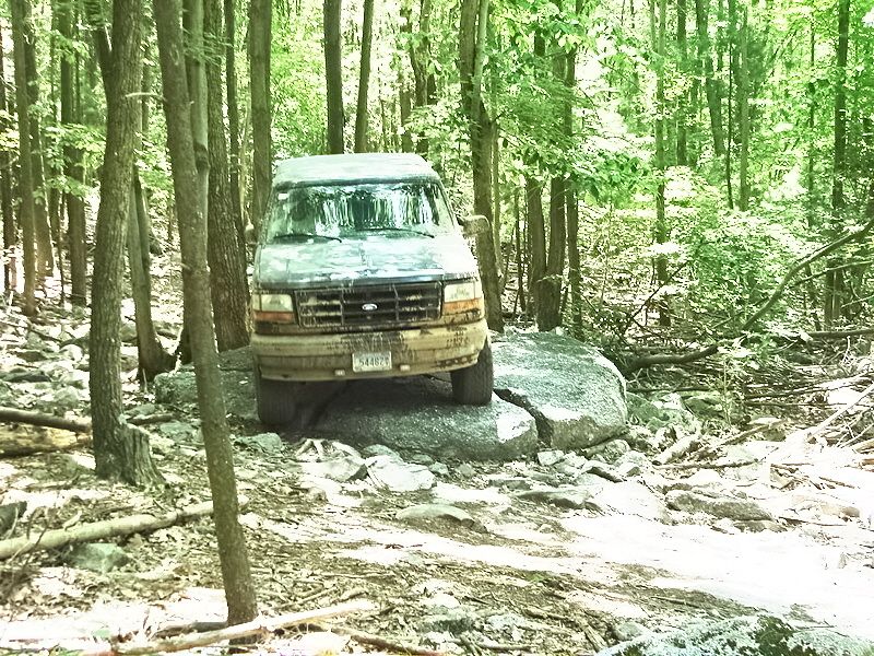 Dark Ford van with front wheels on a boulder on a rocky wooded trail, mud-caked front bumper