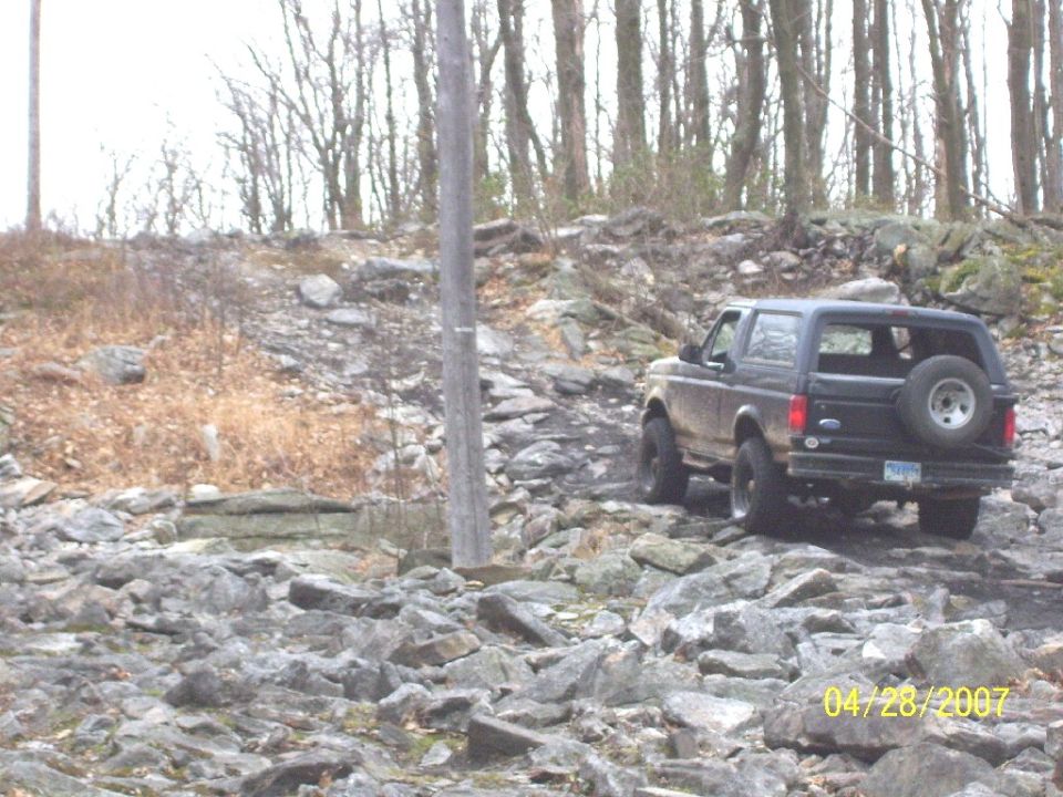 Muddy dark Ford Bronco climbing a rocky trail in leafless woods