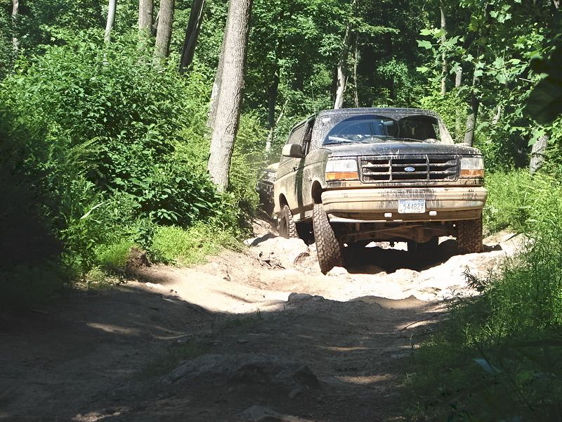 Muddy dark green 1990s Ford Bronco flexing over a rutted dirt trail in the woods