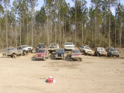 1979 Ford Bronco - A collection of off-road vehicles are gathered on a dirt lot, with a dense forest providing a backdrop.