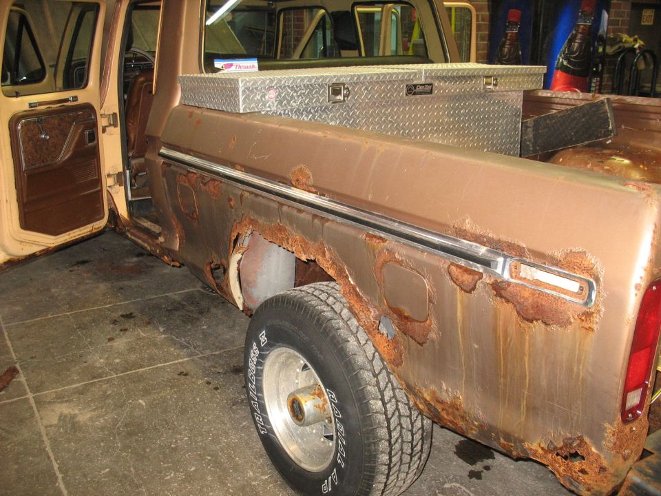 Side view of a heavily rusted pickup truck with the driver's door open. A toolbox is mounted in the bed