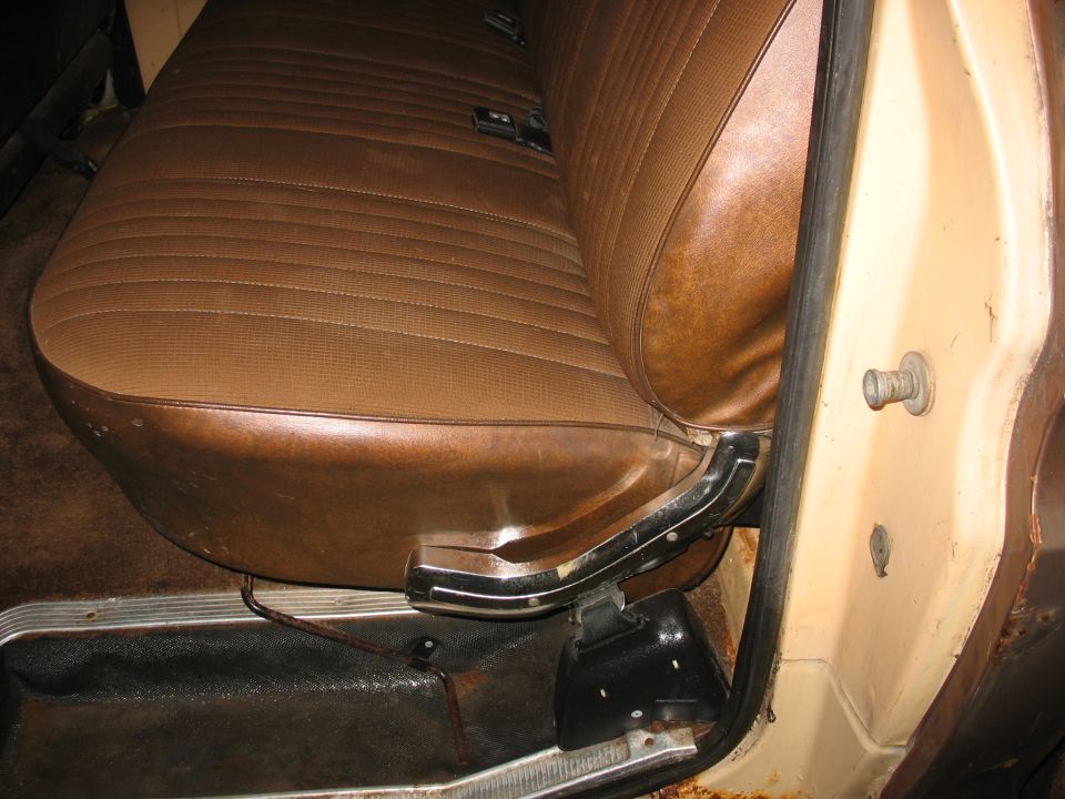 Brown vinyl bench seat with horizontal pleats and a chrome seat adjustment lever. Visible door jamb and floor pan